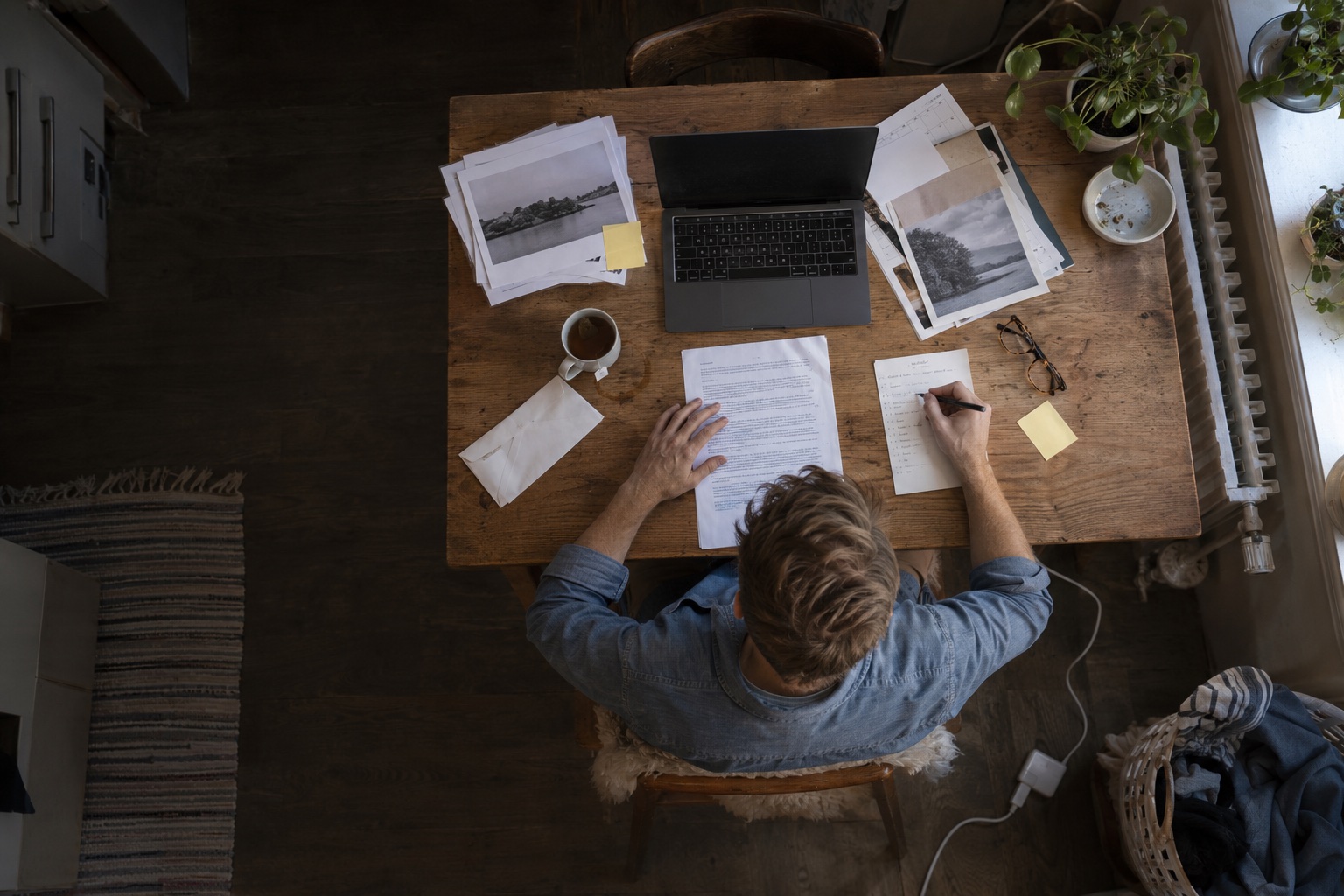 Directly overhead view of a young man in a blue shirt checking a letter and handwritten list at a kitchen-table workspace.