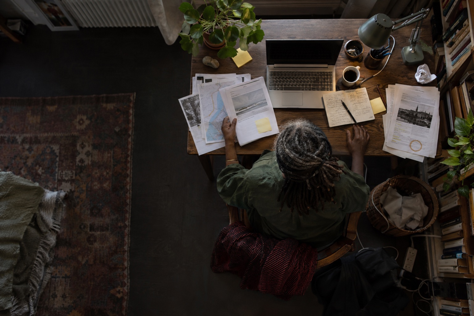 Directly overhead view of an older woman in a green blouse checking reports and source notes in a home office.